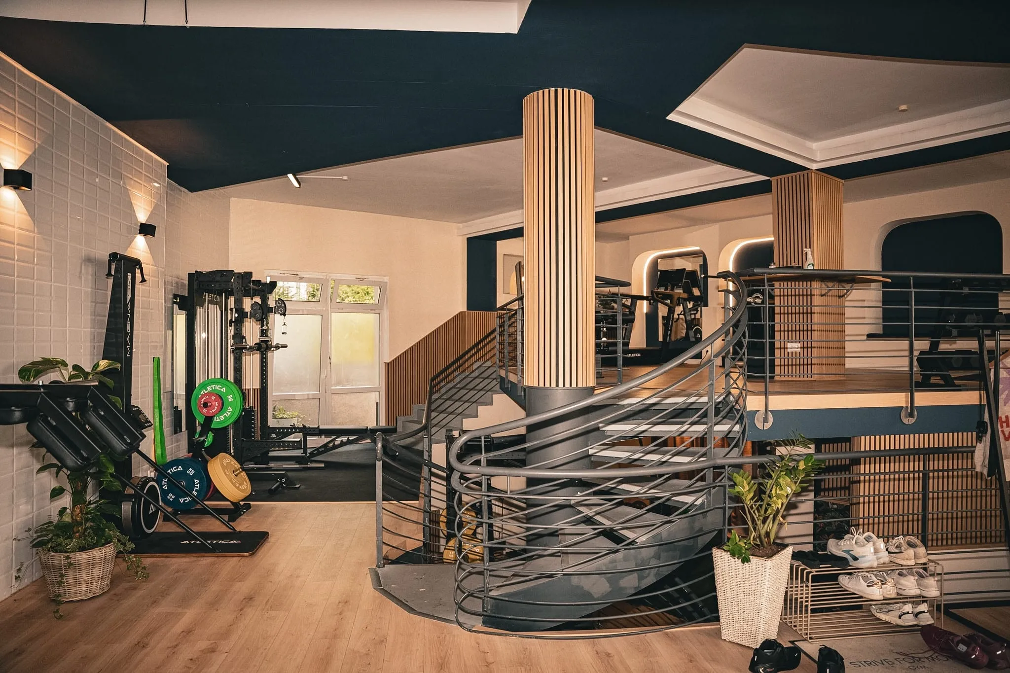 Trainingsfläche des Studios über zwei Ebenen: Rack mit Hantelscheiben, gewendelte Treppe zur Galerie, darüber Cardiogeräte
